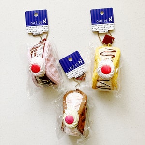 May include: Three miniature cream puff keychain charms with different fillings. Each charm has a blue and white tag with the text "Café de N" and a gold chain attached. The cream puffs are decorated with whipped cream and a red cherry.