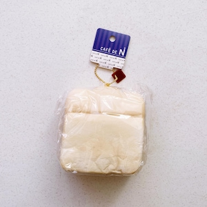 May include: A cream-colored, square-shaped bread product, wrapped in clear plastic. A blue tag with the text "CAFÉ DE N" is attached with a gold chain and a red square charm.