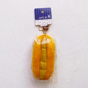 May include: A yellow, foam hot dog toy with a golden-yellow filling, packaged in clear plastic. The toy is attached to a blue tag with the text "CAFÉ DE N". A small gold chain and a brown tag are also attached.