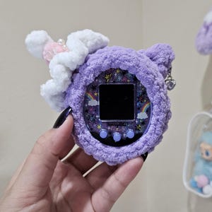 Può includere: Un dispositivo digitale portatile viola e in peluche per animali domestici con una copertura decorativa. La copertura presenta un design a stelle e arcobaleno sullo schermo ed è adornata da una nuvola bianca e accenti rosa. Un piccolo ciondolo pende dal lato.