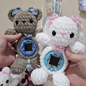 Puede incluir: Dos juguetes de peluche de ganchillo hechos a mano: un oso gris con lazos azules y un conejito blanco con lazos rosas. Cada juguete tiene un dispositivo Tamagotchi en el centro. El oso y el conejito se sostienen frente a una pared.