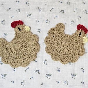 Puede incluir: Dos posavasos hechos a mano de ganchillo con forma de gallina. Los posavasos son de color beige con una cresta roja y una peque&ntilde;a car&uacute;ncula amarilla. Se colocan sobre un fondo blanco con un estampado floral azul.