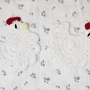 Puede incluir: Dos posavasos blancos de ganchillo con forma de gallina. Cada posavasos tiene una cresta roja, un ojo negro y una car&uacute;ncula amarilla. Los posavasos est&aacute;n sobre un fondo blanco con un estampado floral azul.