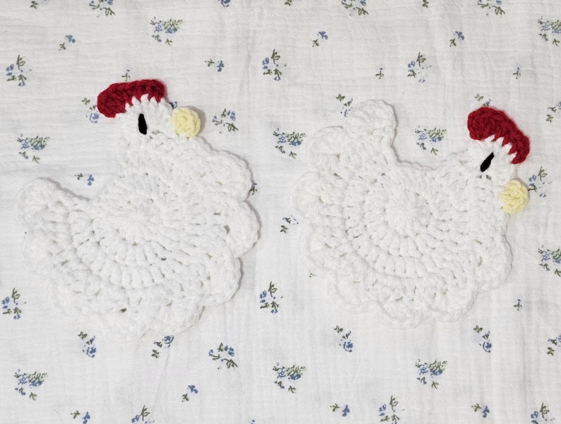 Puede incluir: Dos posavasos blancos de ganchillo con forma de gallina. Cada posavasos tiene una cresta roja, un ojo negro y una car&uacute;ncula amarilla. Los posavasos est&aacute;n sobre un fondo blanco con un estampado floral azul.