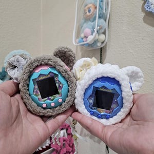 May include: Two Tamagotchi virtual pet devices, one in a gray crocheted case with pink accents and the other in a white crocheted case. Both have bear ear details and floral embellishments. The screens display a digital interface.