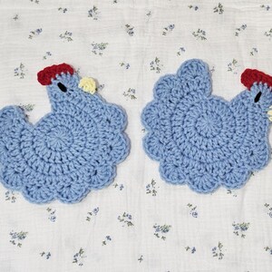 Puede incluir: Dos salvamanteles hechos a mano, de ganchillo azul con forma de gallina. Cada salvamanteles tiene una cresta roja, una car&uacute;ncula amarilla y un peque&ntilde;o ojo negro. Est&aacute;n sobre una tela blanca con un delicado estampado floral.