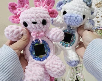 May include: Two crochet axolotl plush toys. One is pink with pink and magenta accents, the other is white with light blue accents. Both have black eyes and a screen in the centre. The pink axolotl has a pink bow and the white one has a blue bow.