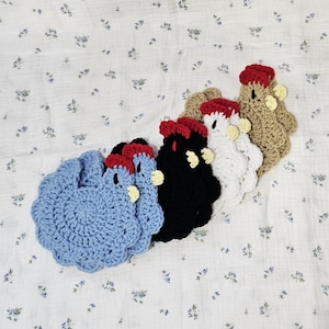 Puede incluir: Un conjunto de cinco posavasos con forma de gallina hechos a ganchillo en varios colores: azul claro, negro, blanco y beige. Cada gallina tiene una cresta roja y un pico y barbillas amarillas. Los posavasos est&aacute;n sobre una superficie blanca con estampado floral.