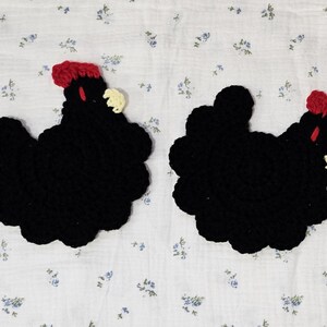 Puede incluir: Dos posavasos hechos a mano en crochet con forma de gallina. Cada posavasos es principalmente negro con una cresta y una barbilla rojas, y un peque&ntilde;o pico color crema. Los posavasos est&aacute;n colocados sobre una tela blanca con un estampado floral azul.