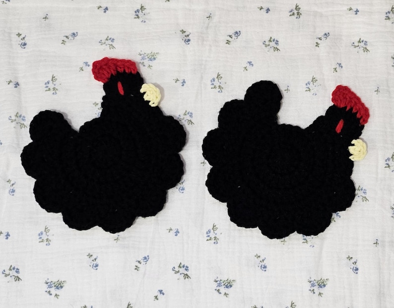 Puede incluir: Dos posavasos hechos a mano en crochet con forma de gallina. Cada posavasos es principalmente negro con una cresta y una barbilla rojas, y un peque&ntilde;o pico color crema. Los posavasos est&aacute;n colocados sobre una tela blanca con un estampado floral azul.
