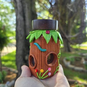 May include: A whimsical, handcrafted jar with a tree trunk design. The brown jar features two round openings, a green leaf and ladybug accent, and a blue caterpillar. Colorful flowers and vines decorate the base. The jar has a dark brown lid.
