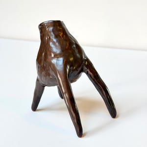 May include: A dark brown ceramic vessel with a glossy, uneven surface and a rounded body. The object features three elongated, tapered legs that extend outward, providing a tripod-like base for the hollow, open-topped structure.