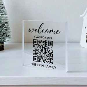 May include: A clear acrylic sign with the words "welcome" and "SCAN FOR WIFI" in black script, along with a QR code. The text "THE ERIN FAMILY" is below the QR code. The sign is on a white surface, with a small Christmas tree and a decorative house.