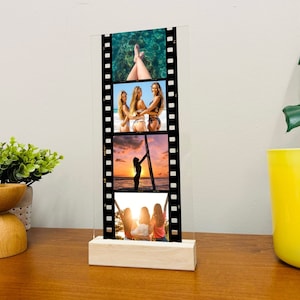 May include: A clear acrylic photo frame with a wood base. The frame features a film strip design with four photos of women in various poses. The photos are of women in swimwear, a woman in silhouette on a beach, and two women in a boat.