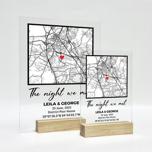 May include: Two clear acrylic map prints with wooden bases. Each print features a black and white map with a red heart marking a location. The text "The night we met" is displayed, along with names, a date, and coordinates.