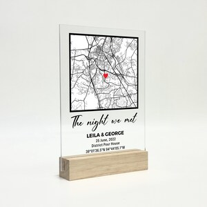 May include: Clear acrylic plaque featuring a black and white map design, a red heart, and the text "The night we met." Includes names, date, and location details. The plaque is set on a light wood base.