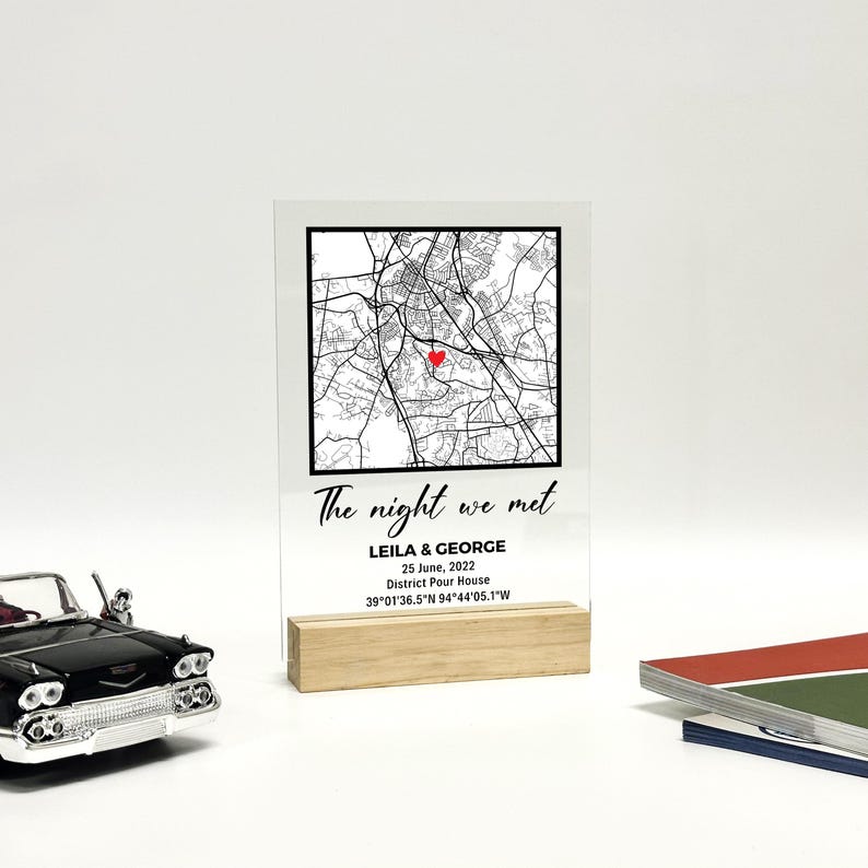 May include: A clear acrylic sign featuring a map graphic with a red heart marking a location. The text "The night we met" is above the names "LEILA & GEORGE" and a date. The sign is supported by a wooden base.