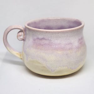 May include: A ceramic mug with a rounded body and a curved handle. The upper portion of the mug features a soft lavender glaze, while the lower portion is finished in a creamy off-white glaze. The two colors blend together in a drip-like pattern.