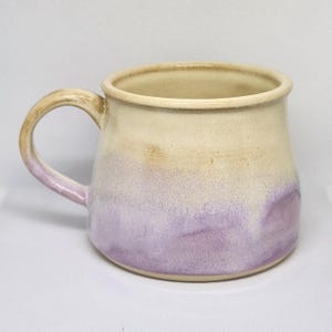 May include: A ceramic mug featuring a two-tone glaze with a creamy off-white upper section and a soft lavender purple lower section. The mug has a rounded body, a wide rim, and a curved handle that transitions from a tan color to a light purple.