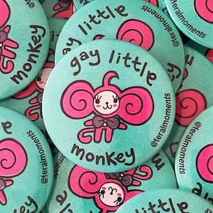 May include: A collection of teal-colored round buttons. Each button features a pink cartoon monkey with the words "gay little monkey" in black text. The buttons are scattered, with some overlapping.