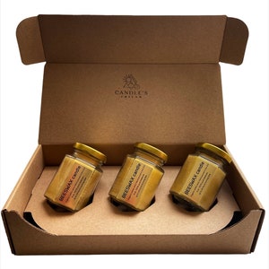 May include: Three glass jars filled with beeswax candles in a brown cardboard box. The jars have gold lids and labels that say "BEESWAX candle" with a description of the candle. The box has a logo that says "CANDLE'S FRIEND".