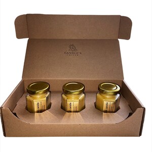 May include: Three small glass jars with gold lids containing yellow wax candles. The jars are in a brown cardboard box with the words "CANDLE'S" printed on the inside.