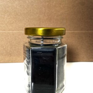 May include: A glass jar with a gold lid filled with black wax. The jar is hexagonal in shape.