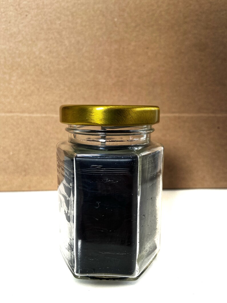 May include: A glass jar with a gold lid filled with black wax. The jar is hexagonal in shape.