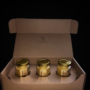 May include: A cardboard box with three small glass jars containing yellow wax candles. The jars have gold lids and labels that say "Soy candle" and "Hand poured in the USA". The box has a logo that says "Candle's Ritual".