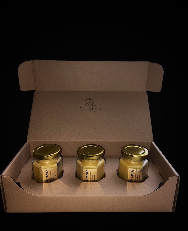 May include: A cardboard box with three small glass jars containing yellow wax candles. The jars have gold lids and labels that say "Soy candle" and "Hand poured in the USA". The box has a logo that says "Candle's Ritual".