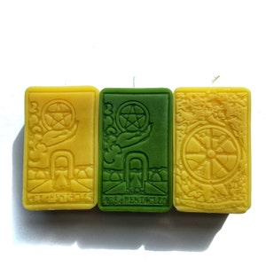 May include: Three tarot card-themed candles, one green and two yellow. The green candle is the Ace of Pentacles and the yellow candles are the 3 of Pentacles and the Wheel of Fortune.