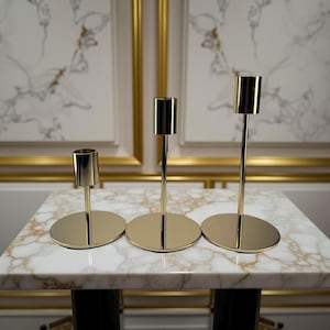May include: Three gold-colored candle holders of varying heights. Each holder has a circular base and a slender stem leading to a cylindrical cup. The holders are arranged on a marble-patterned surface.