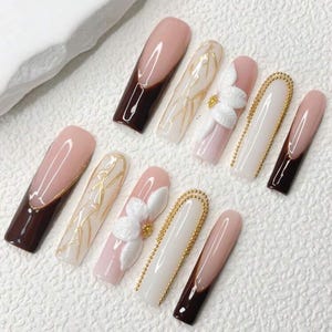 May include: A set of ten long, artificial nails. The nails feature a variety of designs, including a French tip with a dark brown color, gold accents, white floral embellishments, and gold bead detailing. The nails have a glossy finish.