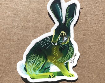 May include: A sticker featuring a stylized illustration of a hare in shades of green and yellow. The hare is depicted in a seated position with long ears and a white outline. The artist's signature and date, "N. John '25", are visible.