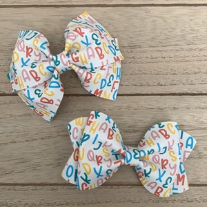 Può includere: Due fiocchi per capelli bianchi con una stampa alfabetica colorata. I fiocchi sono fatti di tessuto e hanno uno sfondo bianco con lettere colorate.