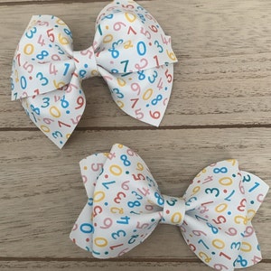 Può includere: Due fiocchi per capelli bianchi con una stampa di numeri colorati. I fiocchi sono fatti di tessuto e hanno uno sfondo bianco con numeri colorati stampati su di essi. I numeri sono di diversi colori, tra cui rosso, blu, giallo e verde.