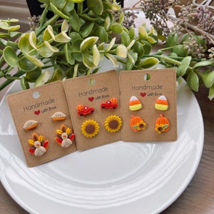 May include: Three sets of handmade earrings with fall-themed designs. The first set features a pumpkin, a sunflower, and a candy corn. The second set features a sunflower, a car, and a pumpkin. The third set features a turkey, a sunflower, and a pie.