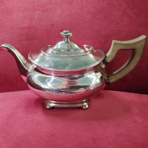 Puede incluir: Una tetera plateada con asa de madera y tapa a juego. La tetera tiene un pico curvo y se asienta sobre una base cuadrada. El asa es de madera clara.