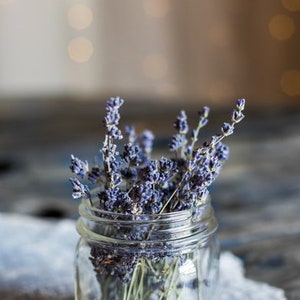 Op de afbeelding: Een glazen pot gevuld met gedroogde lavendelbloemen. De pot staat op een wit kantonderzetter.