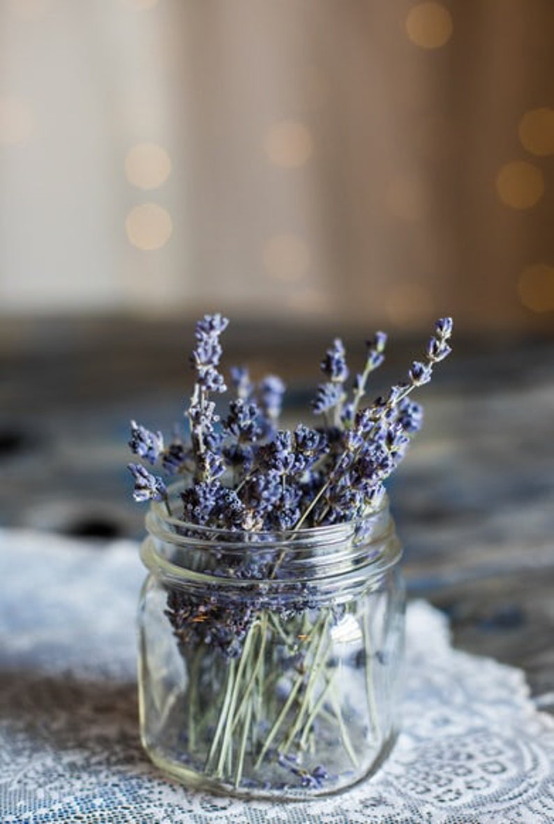 Op de afbeelding: Een glazen pot gevuld met gedroogde lavendelbloemen. De pot staat op een wit kantonderzetter.