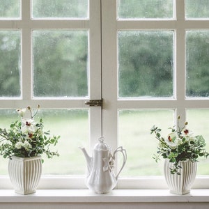 May include: A white windowsill with three white ceramic items. Two white ceramic vases with green leafy plants and white flowers. A white ceramic teapot with a floral design.