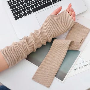 Op de afbeelding: Een paar beige gebreide armwarmers. De armwarmers zijn geribbeld en hebben een lang, vloeiend ontwerp. De armwarmers liggen op een wit oppervlak.