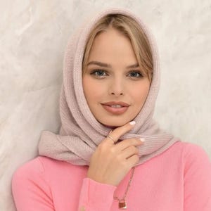 May include: A woman wearing a light pink knitted scarf with a hood. The scarf is draped around her neck and shoulders. She is wearing a pink sweater and has a gold necklace with a padlock pendant.