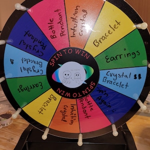 May include: A colorful wheel with a black background and white spokes. The wheel is divided into 10 sections, each with a different color and a prize written on it. The prizes include: "Crystal Bracelet", "Earrings", "Crystal Bracelet", "Spin Again", "Bottle Pendant", "Intuition Crystal", "Bottle Pendant", "Crystal Bracelet", "Earrings", and "Crystal Bracelet". The wheel is labeled "Spin to Win" and "Raine & Sea".