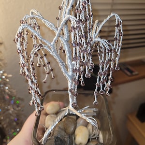 May include: A silver wire tree sculpture with brown glass beads hanging from the branches. The tree is in a clear glass vase with small rocks at the base.