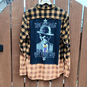 May include: A plaid flannel shirt in shades of yellow and black, with a large black patch on the back featuring a skull wearing a hat and suit. The text on the patch reads "SIGMA ALPHA EPSILON" and "PADDY MURPHY WEEK."