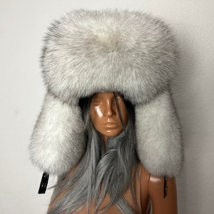 Peut inclure: Un chapeau d'hiver luxueux et duveteux dans les tons de gris et de blanc. Le chapeau présente une grande couronne arrondie et des rabats d'oreilles qui s'étendent vers le bas. Des lanières en cuir noir avec des boucles sont attachées aux rabats d'oreilles. Le chapeau est en fourrure douce et texturée.