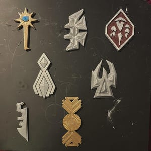 Può includere: Un assortimento di oggetti decorativi geometrici color metallo. Gli oggetti includono una stella, una forma a diamante e altri disegni astratti. Gli oggetti sono nei colori oro, argento e bordeaux. Sono disposti su una superficie scura.