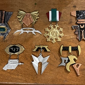 Könnte beinhalten: Acht metallische Broschen in verschiedenen Designs, mit Silber, Gold, Kupfer und Schwarz. Einige haben geometrische Formen, andere aufwendigere Elemente. Die Broschen werden auf einer Holzoberfläche präsentiert und zeigen eine Reihe von Stilen und Farben.