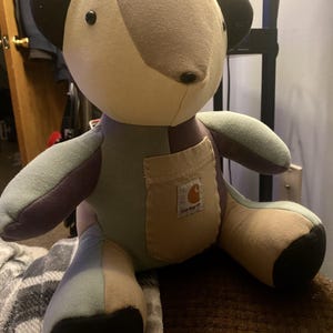 May include: A patchwork teddy bear in various shades of beige, grey, and blue. The bear has black button eyes and a small pocket with the Carhartt logo. The bear is a handmade item.